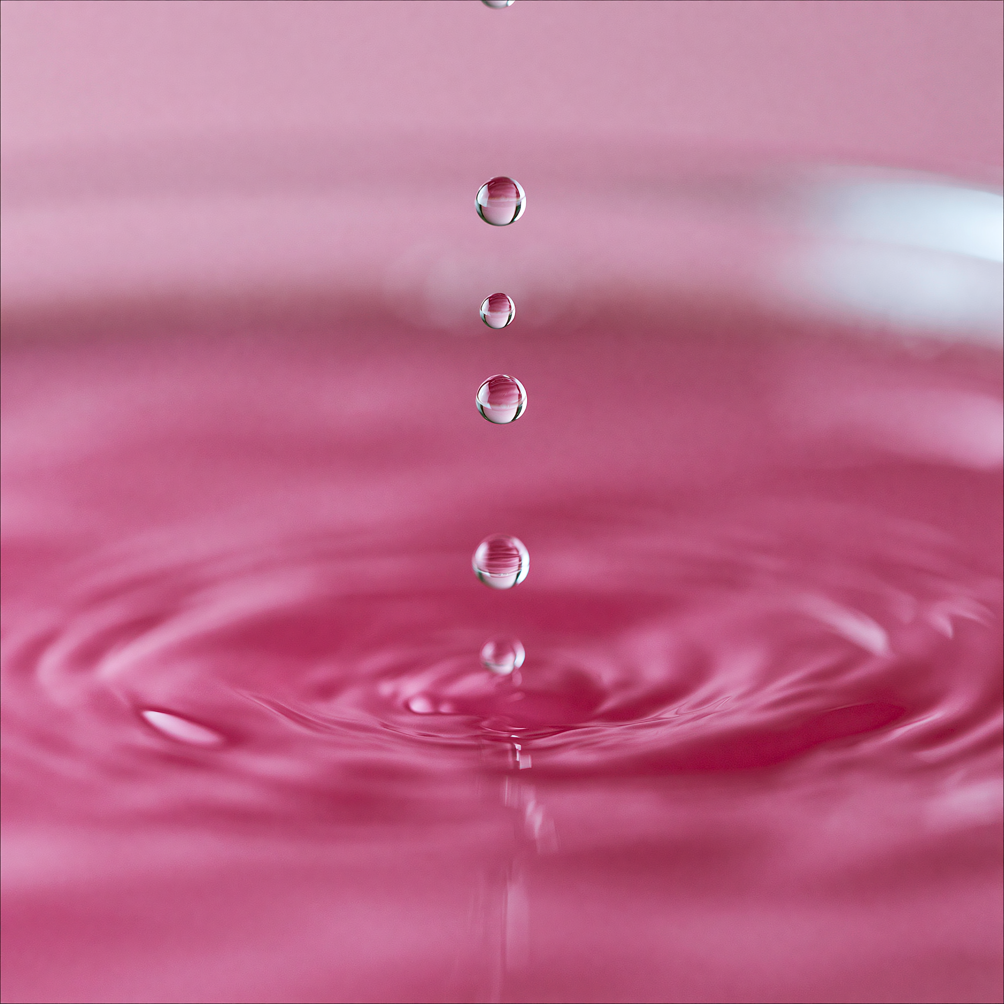 A close-up photograph of pink liquid with a vertical column of water droplets falling into the surface, creating concentric ripples.